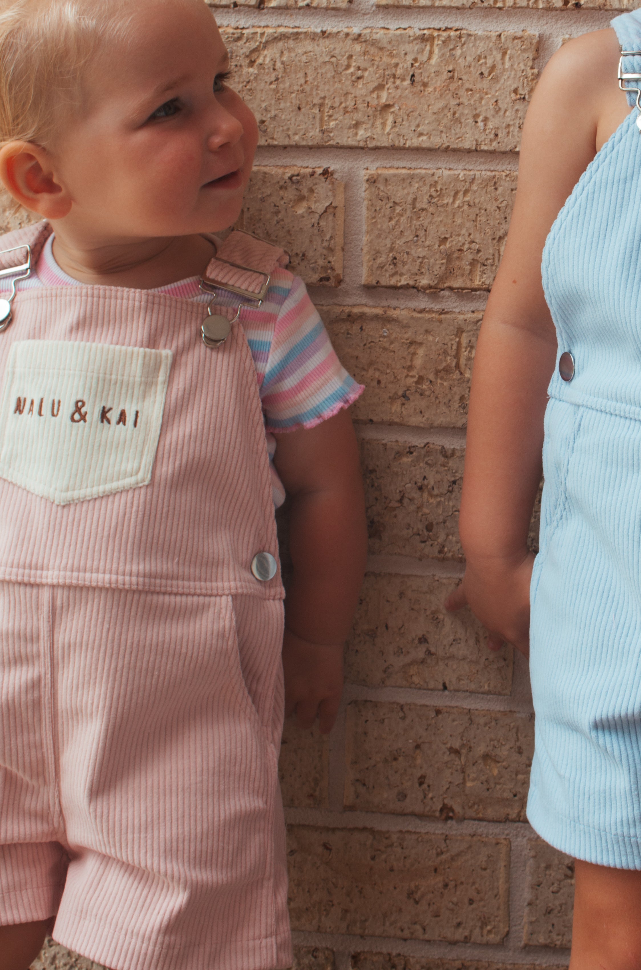 Two children wearing matching overalls with a brand label against a brick wall.
